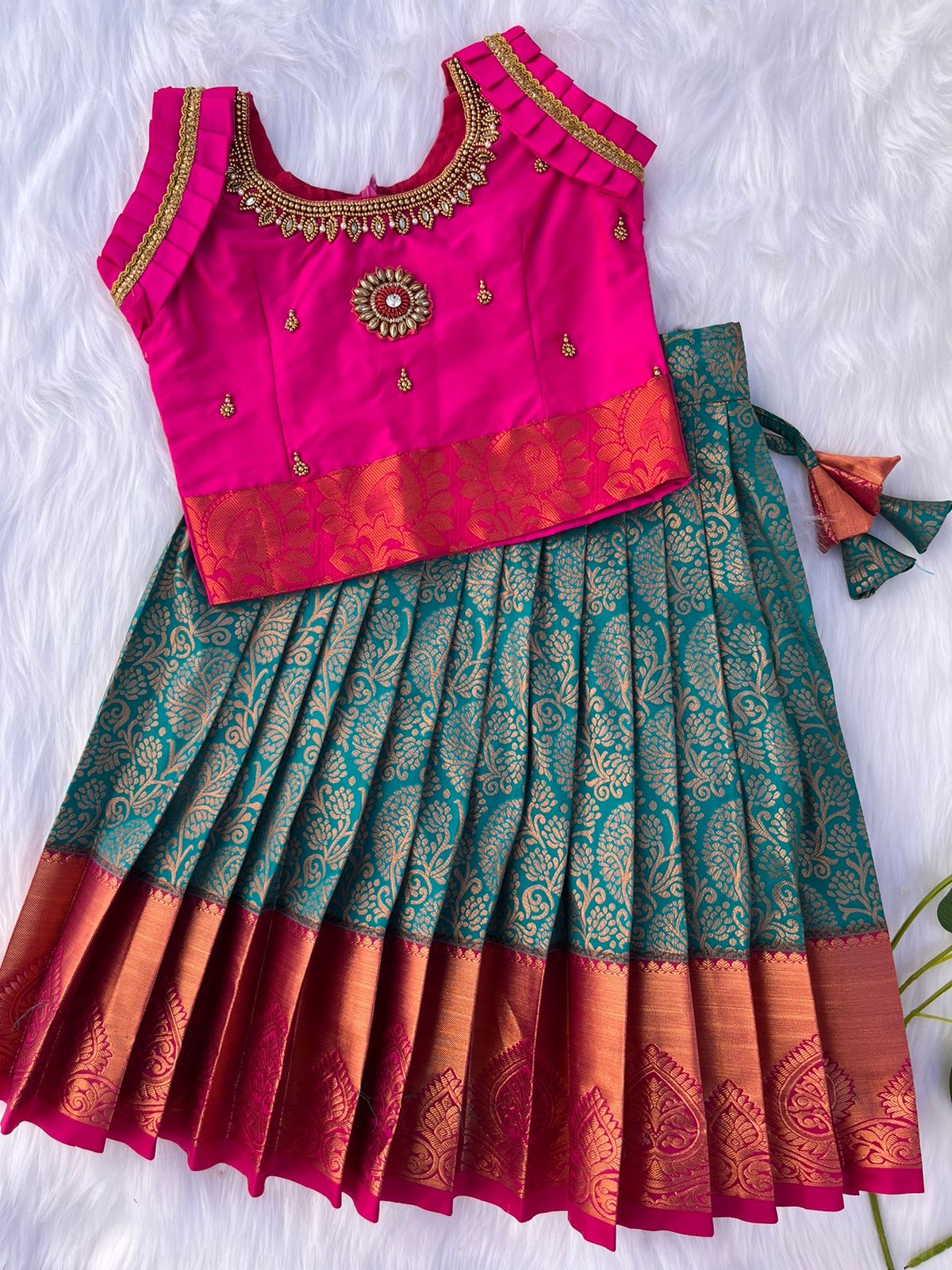 A pink and green semi-silk lehenga with a sleeveless blouse, featuring gold embroidery and zari work. The lehenga has a pleated design with brocade-style patterns and a contrasting pink and orange border.